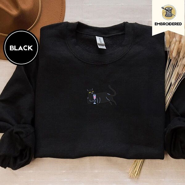 Black crewneck sweatshirt with a brown felt hat and dried bunny tail grass, embroidered with a black cat lounging with a glass of pink wine in the center of the chest