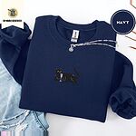 Navy crewneck sweatshirt folded with ripped jeans and a silver chain necklace, embroidered with a black cat lounging with a glass of pink wine in the center of the chest
