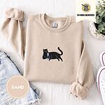 Sand crewneck sweatshirt folded with denim shorts, white sneakers and a potted plant, embroidered with a black cat lounging with a glass of pink wine in the center of the chest