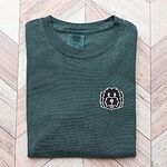 Blue Spruce T-shirt laid flat on a herringbone wood floor, embroidered with a black cartoon doodle face with a pale grey outline and a pink tongue on the left chest