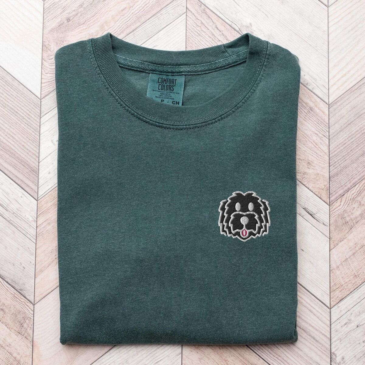 Black Goldendoodle Shirt – Blue Spruce Blue Spruce T-shirt laid flat on a herringbone wood floor, embroidered with a black cartoon doodle face with a pale grey outline and a pink tongue on the left chest