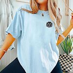 Woman beside a plant in a Chambray T-shirt embroidered with a black cartoon doodle face with a pale grey outline and a pink tongue on the left chest