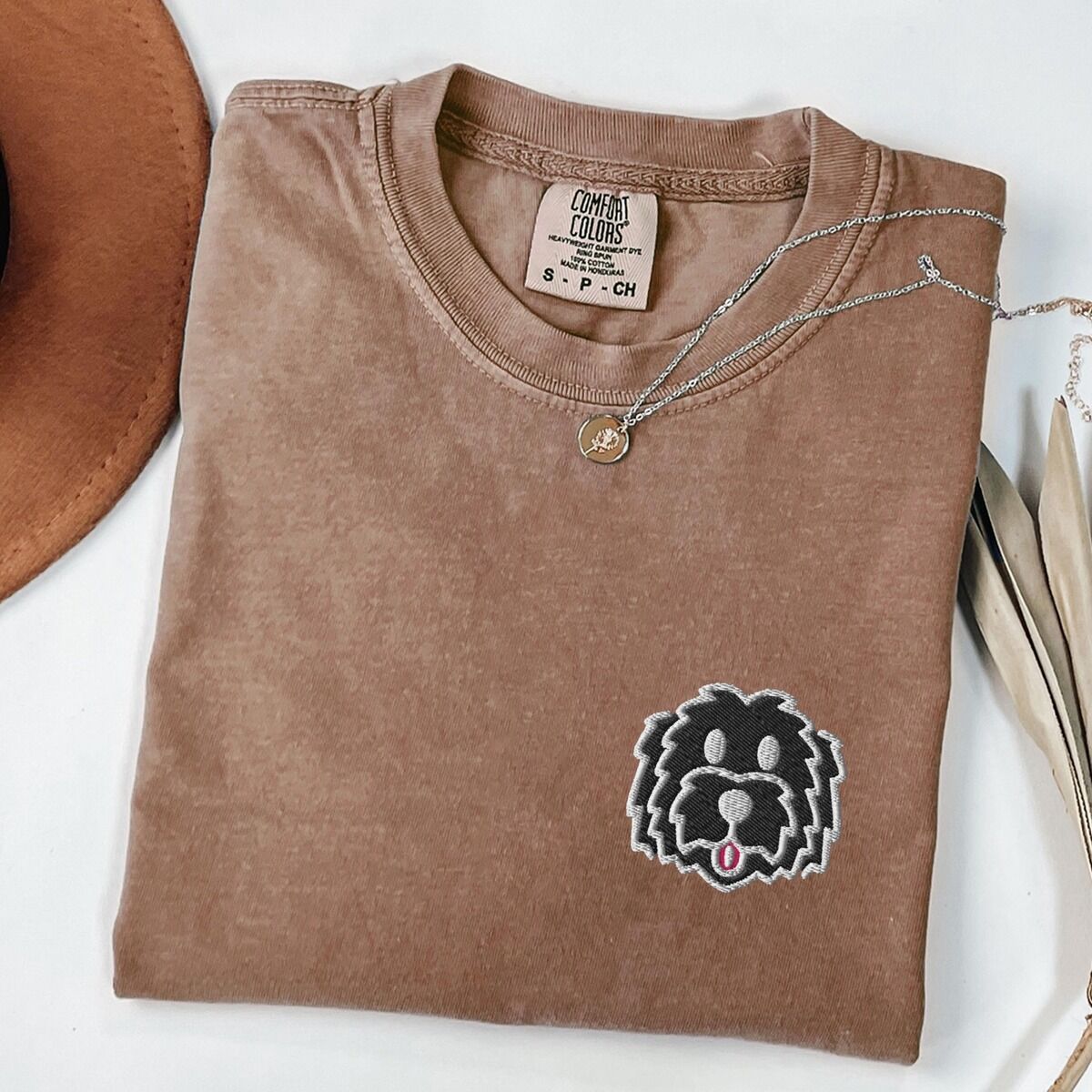 Black Goldendoodle Shirt – Espresso Espresso T-shirt laid flat with a tan hat, a gold necklace and dried palm leaves, embroidered with a black cartoon doodle face with a pale grey outline and a pink tongue on the left chest
