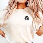 Woman with long wavy hair in an Ivory T-shirt embroidered with a black cartoon doodle face with a pale grey outline and a pink tongue on the left chest
