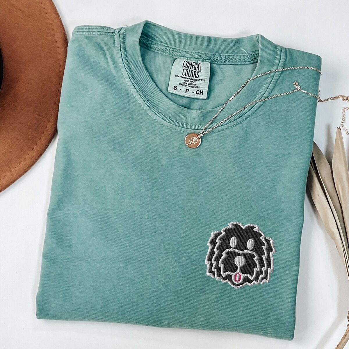 Black Goldendoodle Shirt – Light Green Light Green T-shirt laid flat with a tan hat, a gold necklace and dried palm leaves, embroidered with a black cartoon doodle face with a pale grey outline and a pink tongue on the left chest