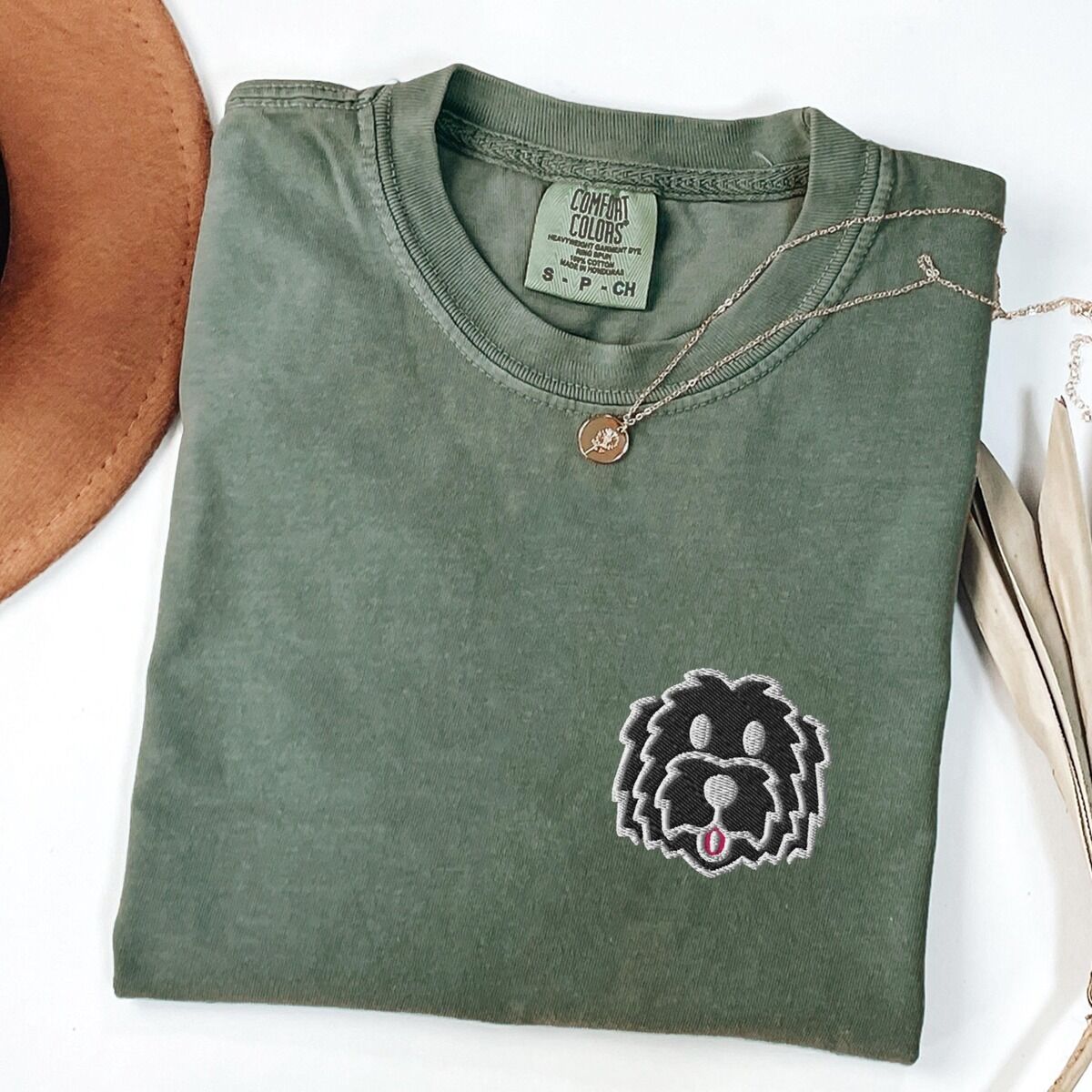 Black Goldendoodle Shirt – Moss Moss T-shirt laid flat with a tan hat, a gold necklace and dried palm leaves, embroidered with a black cartoon doodle face with a pale grey outline and a pink tongue on the left chest