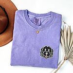 Violet T-shirt laid flat with a tan hat, a gold necklace and dried palm leaves, embroidered with a black cartoon doodle face with a pale grey outline and a pink tongue on the left chest