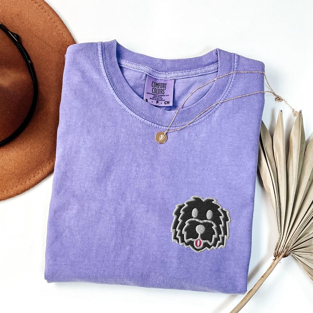 Black Goldendoodle Shirt – Violet Violet T-shirt laid flat with a tan hat, a gold necklace and dried palm leaves, embroidered with a black cartoon doodle face with a pale grey outline and a pink tongue on the left chest
