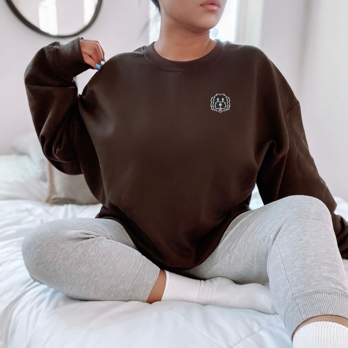 Black Goldendoodle Sweatshirt – Dark Chocolate Woman sitting on a bed in a Dark Chocolate crewneck sweatshirt embroidered with a black cartoon doodle face with a pale grey outline and a pink tongue on the left chest
