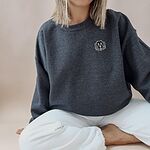 Woman sitting cross-legged in a Dark Heather crewneck sweatshirt embroidered with a black cartoon doodle face with a pale grey outline and a pink tongue on the left chest
