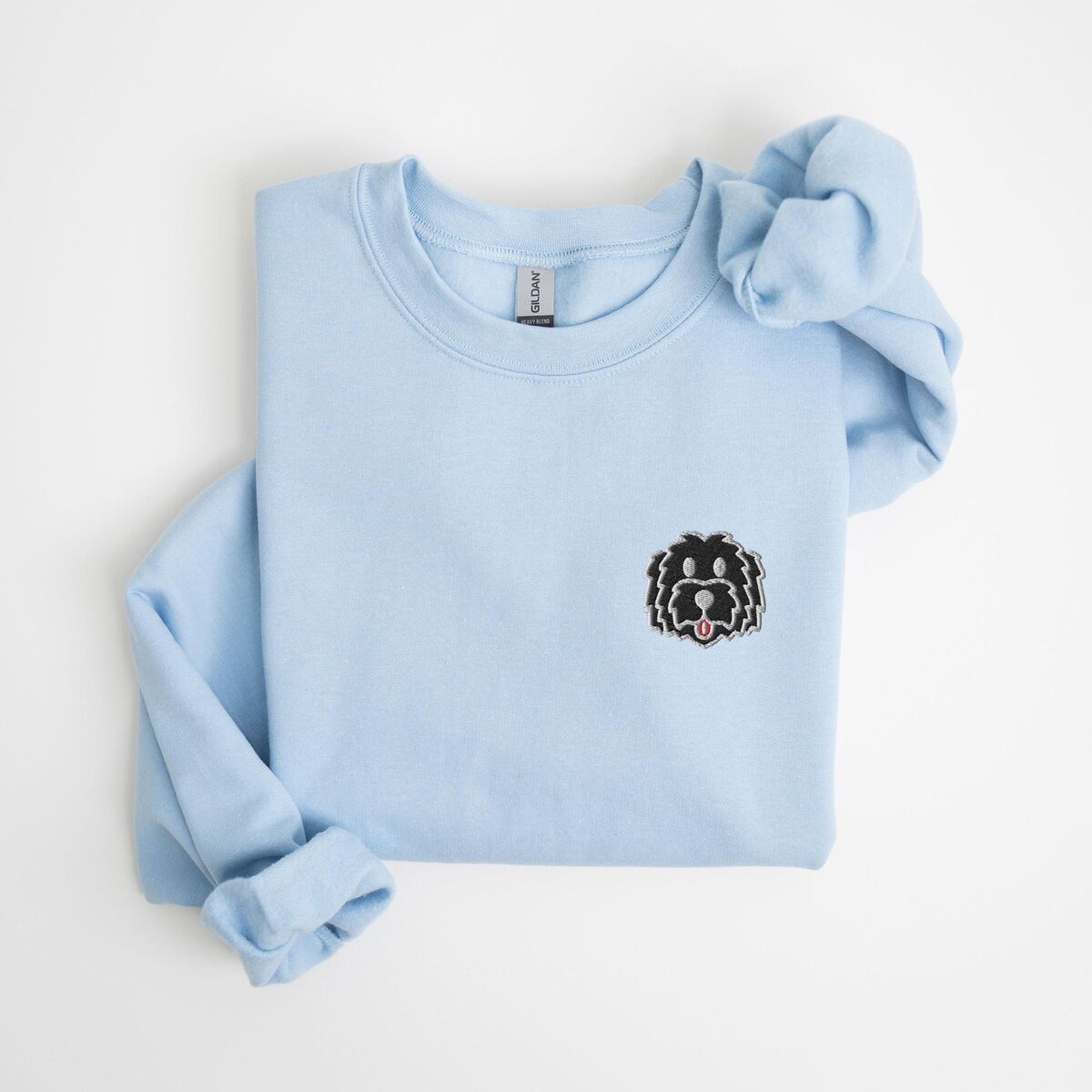 Black Goldendoodle Sweatshirt - Light Blue Light Blue crewneck sweatshirt laid flat on a white background, embroidered with a black cartoon doodle face with a pale grey outline and a pink tongue on the left chest