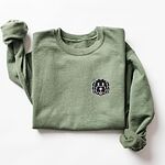 Military Green crewneck sweatshirt laid flat on a white background, embroidered with a black cartoon doodle face with a pale grey outline and a pink tongue on the left chest