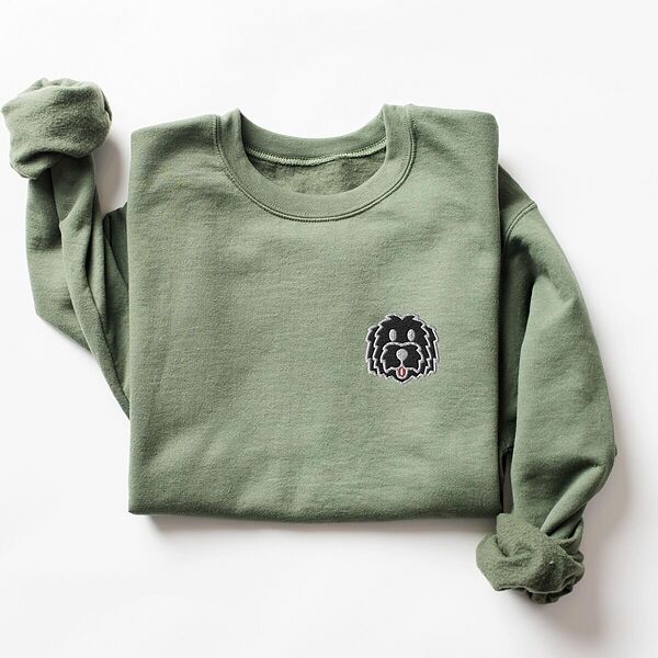 Military Green crewneck sweatshirt laid flat on a white background, embroidered with a black cartoon doodle face with a pale grey outline and a pink tongue on the left chest