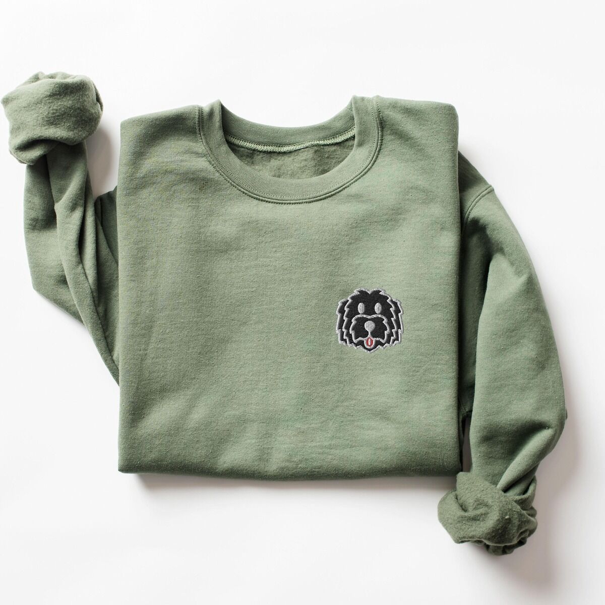 Black Goldendoodle Sweatshirt – Military Green Military Green crewneck sweatshirt laid flat on a white background, embroidered with a black cartoon doodle face with a pale grey outline and a pink tongue on the left chest