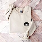 Sand crewneck sweatshirt laid flat on a herringbone wood floor, embroidered with a black cartoon doodle face with a pale grey outline and a pink tongue on the left chest