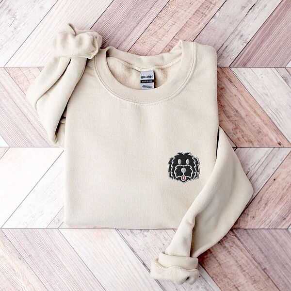 Sand crewneck sweatshirt laid flat on a herringbone wood floor, embroidered with a black cartoon doodle face with a pale grey outline and a pink tongue on the left chest