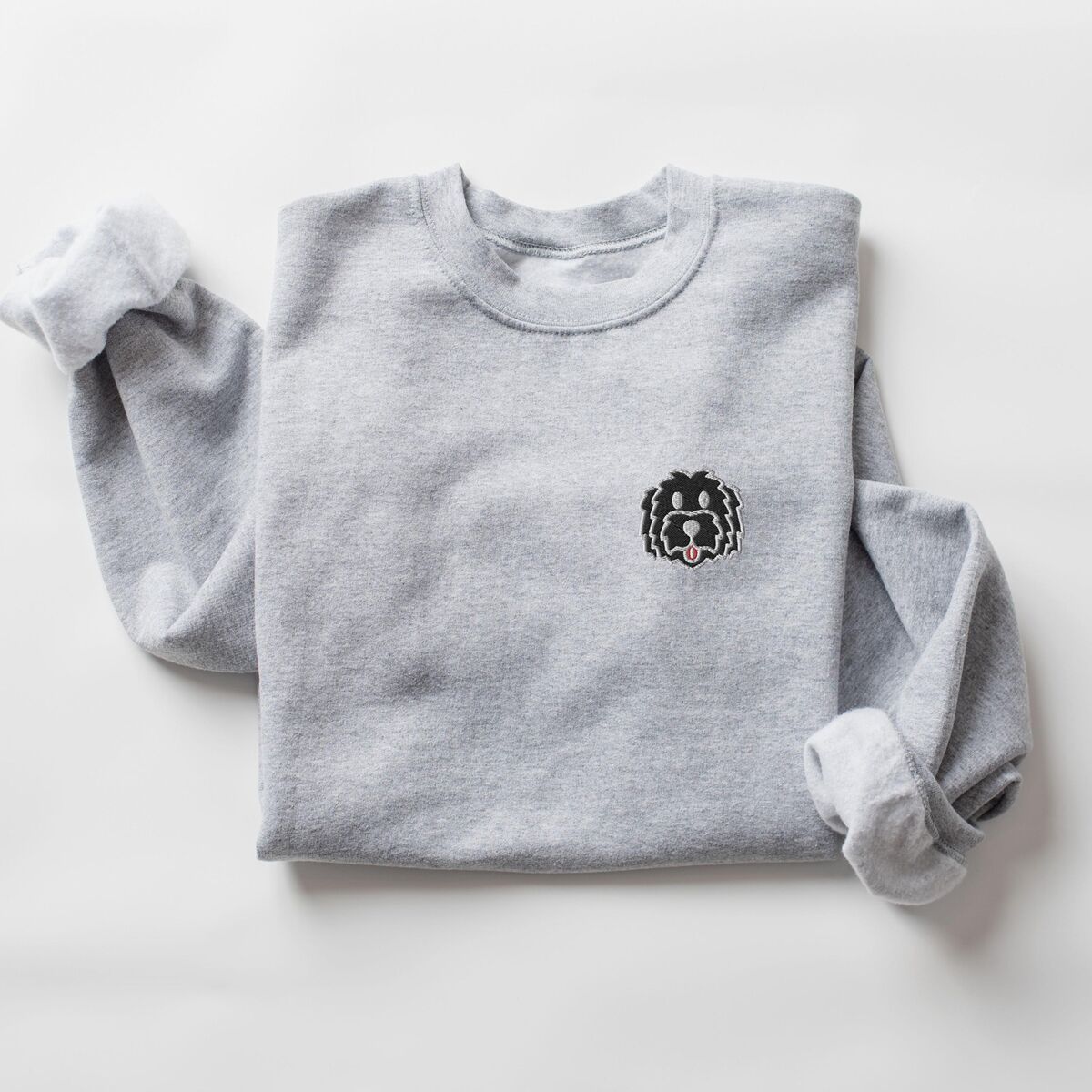 Black Goldendoodle Sweatshirt - Sport Grey Sport Grey crewneck sweatshirt laid flat on a white background, embroidered with a black cartoon doodle face with a pale grey outline and a pink tongue on the left chest