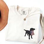 Ivory T-shirt folded beside a hat with a black lab silhouette carrying an American flag on the left chest