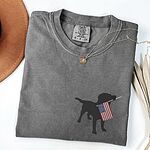 Pepper T-shirt laid flat beside a hat, embroidered with a black lab holding a USA flag on the left chest