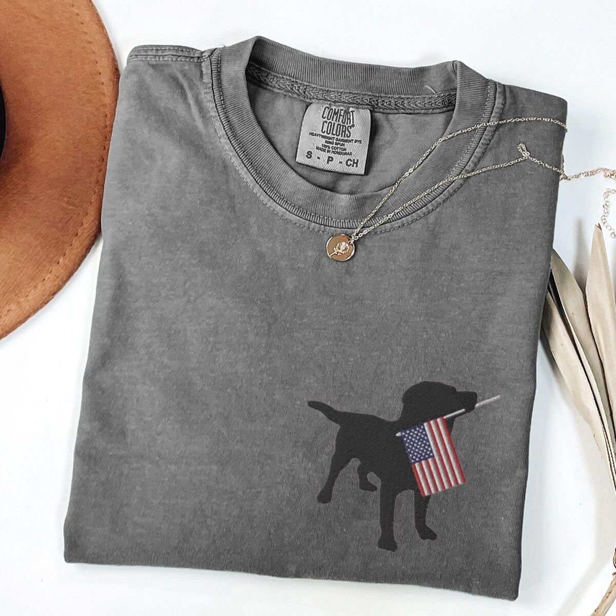 Black Lab American Flag Shirt – Pepper Pepper T-shirt laid flat beside a hat, embroidered with a black lab holding a USA flag on the left chest