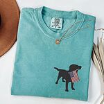 Seafoam T-shirt laid flat beside a hat, embroidered with a black lab holding an American flag on the left chest
