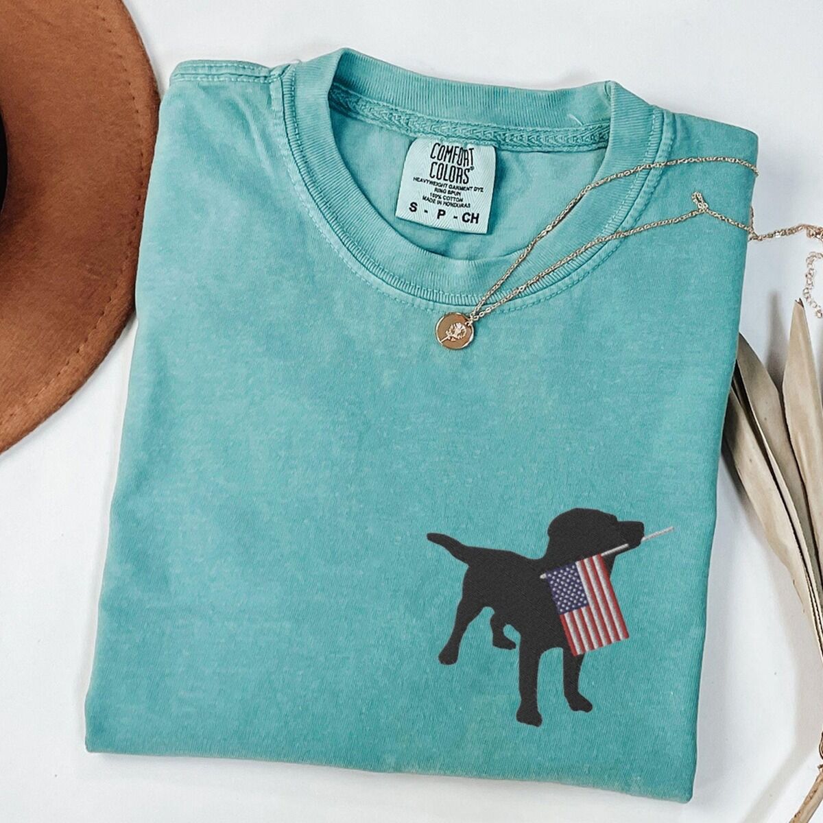 Black Lab American Flag Shirt – Seafoam Seafoam T-shirt laid flat beside a hat, embroidered with a black lab holding an American flag on the left chest