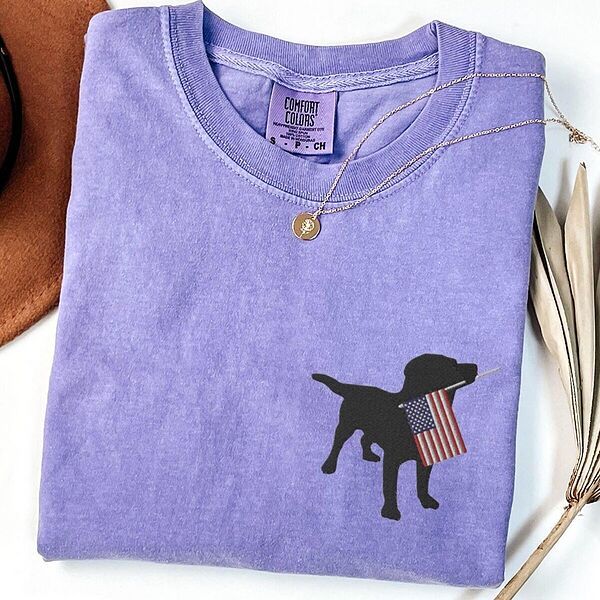 Violet T-shirt laid flat with a necklace, a black Labrador holding a stars-and-stripes flag on the left chest