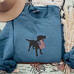 Indigo Blue crewneck sweatshirt beside a hat and dried grass, embroidered with a black lab silhouette carrying an American flag