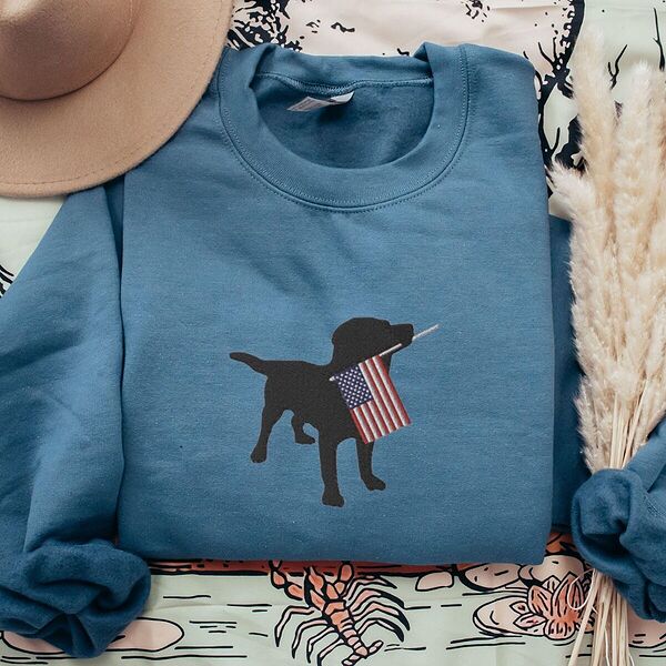 Indigo Blue crewneck sweatshirt beside a hat and dried grass, embroidered with a black lab silhouette carrying an American flag