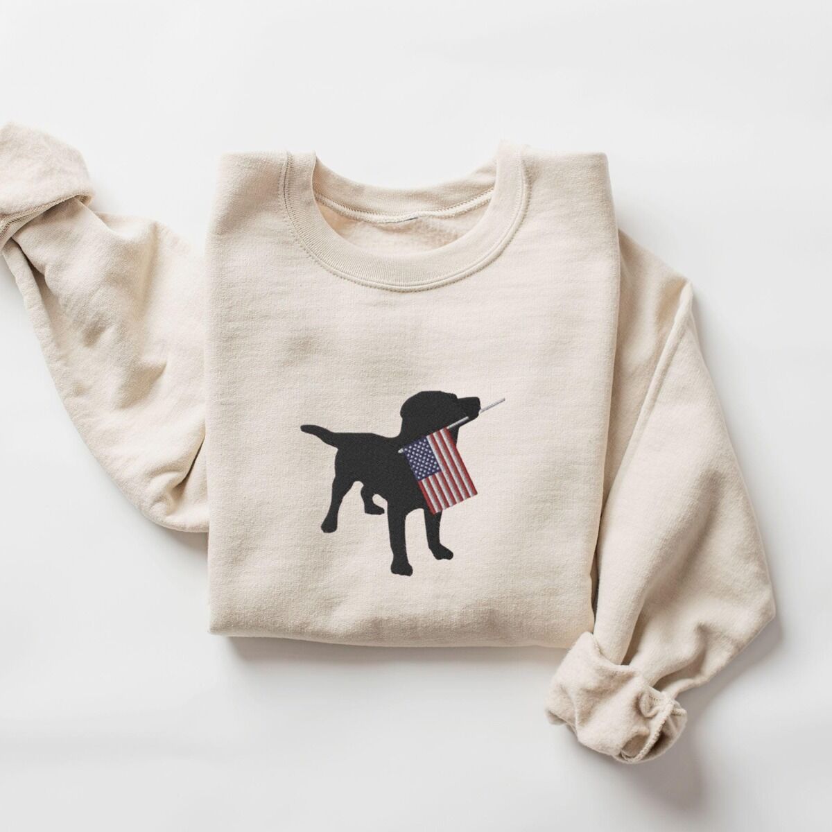 Black Lab American Flag Sweatshirt – Sand Sand crewneck sweatshirt laid flat with a rolled sleeve, embroidered with a black lab carrying a USA flag