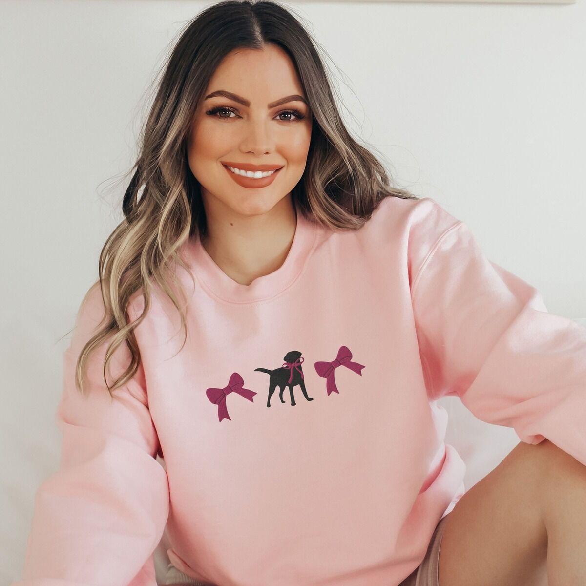 Black Lab Bow Sweatshirt - worn in pink Smiling woman in a Light Pink crewneck sweatshirt embroidered with a black Labrador silhouette with a ribbon bow at its neck between two large raspberry pink bows across the chest