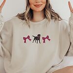 Woman making peace signs in a Sand crewneck sweatshirt embroidered with a black Labrador silhouette with a ribbon bow at its neck between two large raspberry pink bows across the chest