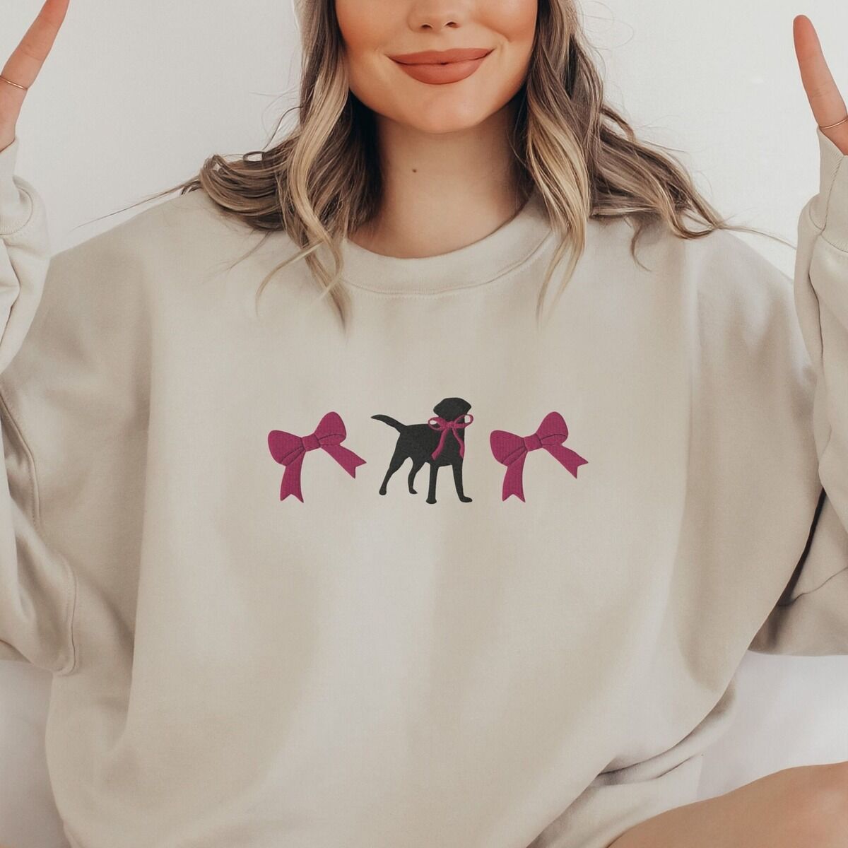 Black Lab Bow Sweatshirt - worn in sand Woman making peace signs in a Sand crewneck sweatshirt embroidered with a black Labrador silhouette with a ribbon bow at its neck between two large raspberry pink bows across the chest
