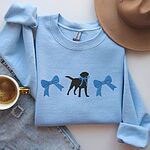 Light Blue crewneck sweatshirt laid flat with jeans, a hat and coffee, embroidered with a black Labrador silhouette with a ribbon bow at its neck between two large blue bows across the chest