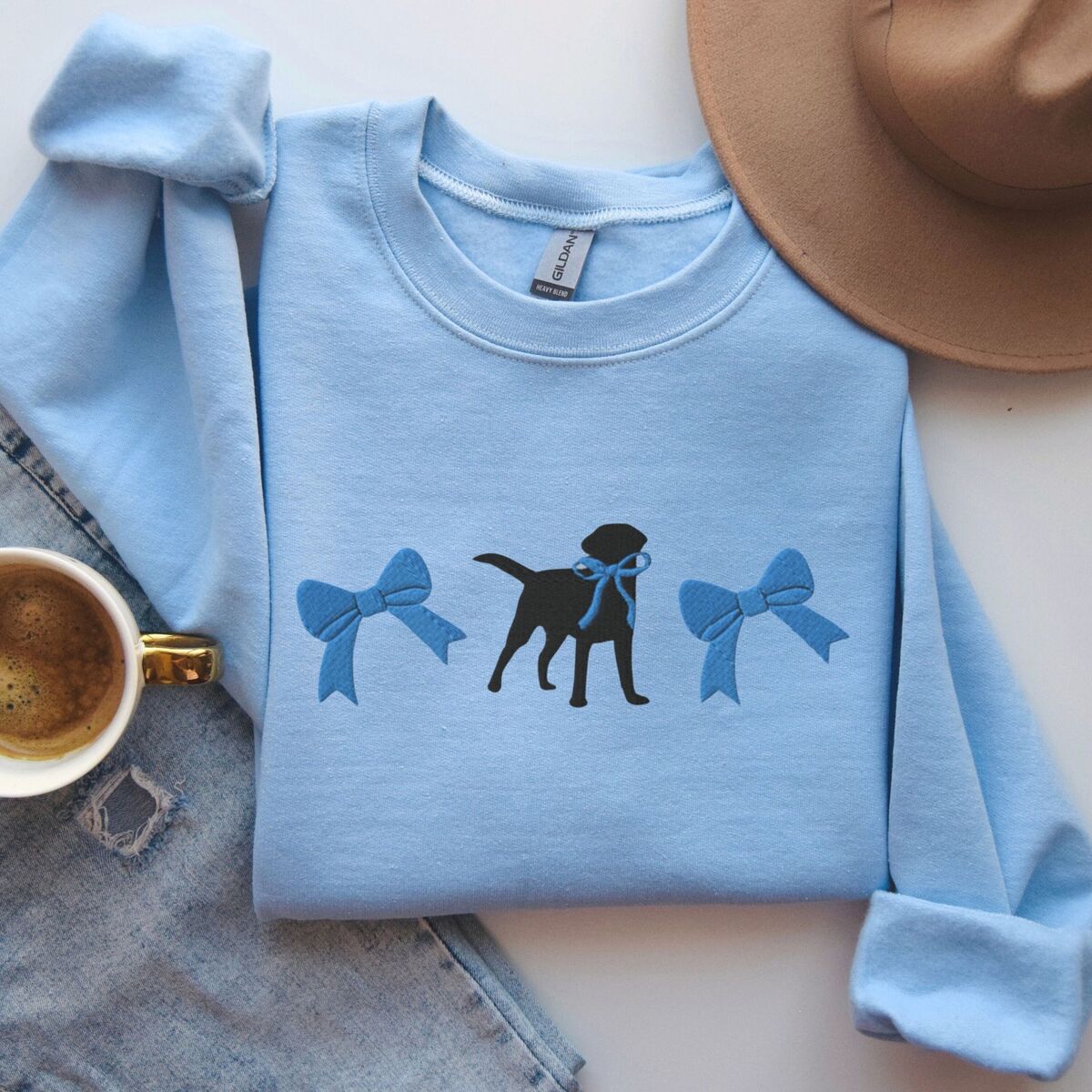 Black Lab Bow Sweatshirt - Light Blue Light Blue crewneck sweatshirt laid flat with jeans, a hat and coffee, embroidered with a black Labrador silhouette with a ribbon bow at its neck between two large blue bows across the chest