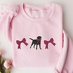 Light Pink crewneck sweatshirt laid flat with pink roses, embroidered with a black Labrador silhouette with a ribbon bow at its neck between two large raspberry pink bows across the chest