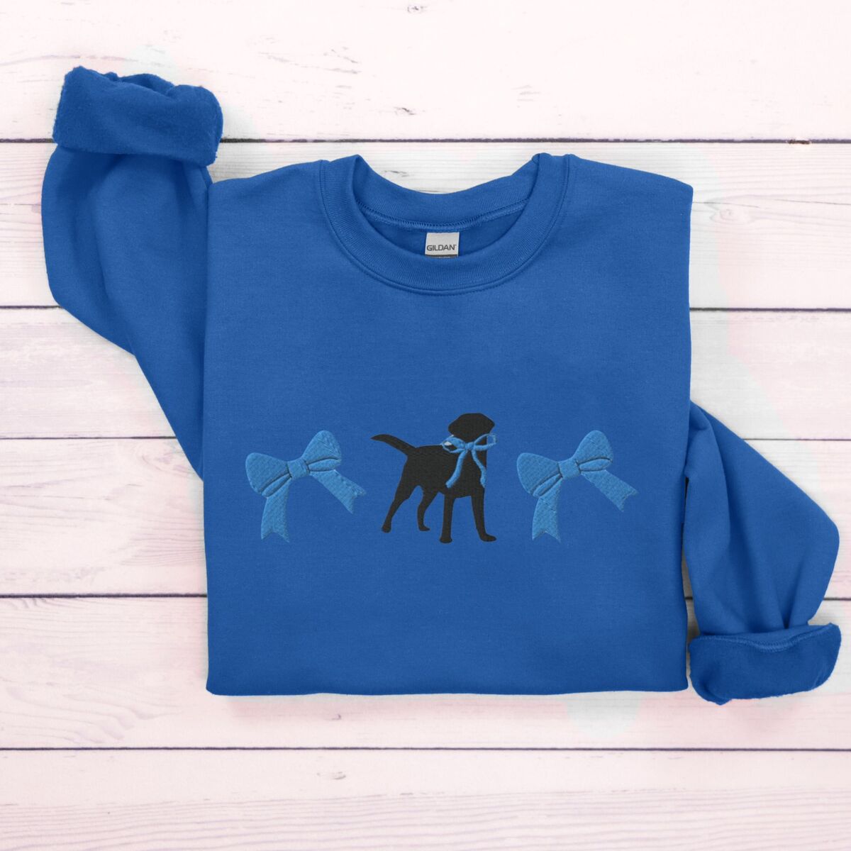 Black Lab Bow Sweatshirt - Royal Royal crewneck sweatshirt laid flat on white wood, embroidered with a black Labrador silhouette with a ribbon bow at its neck between two large blue bows across the chest