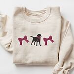 Sand crewneck sweatshirt laid flat on white, embroidered with a black Labrador silhouette with a ribbon bow at its neck between two large raspberry pink bows across the chest
