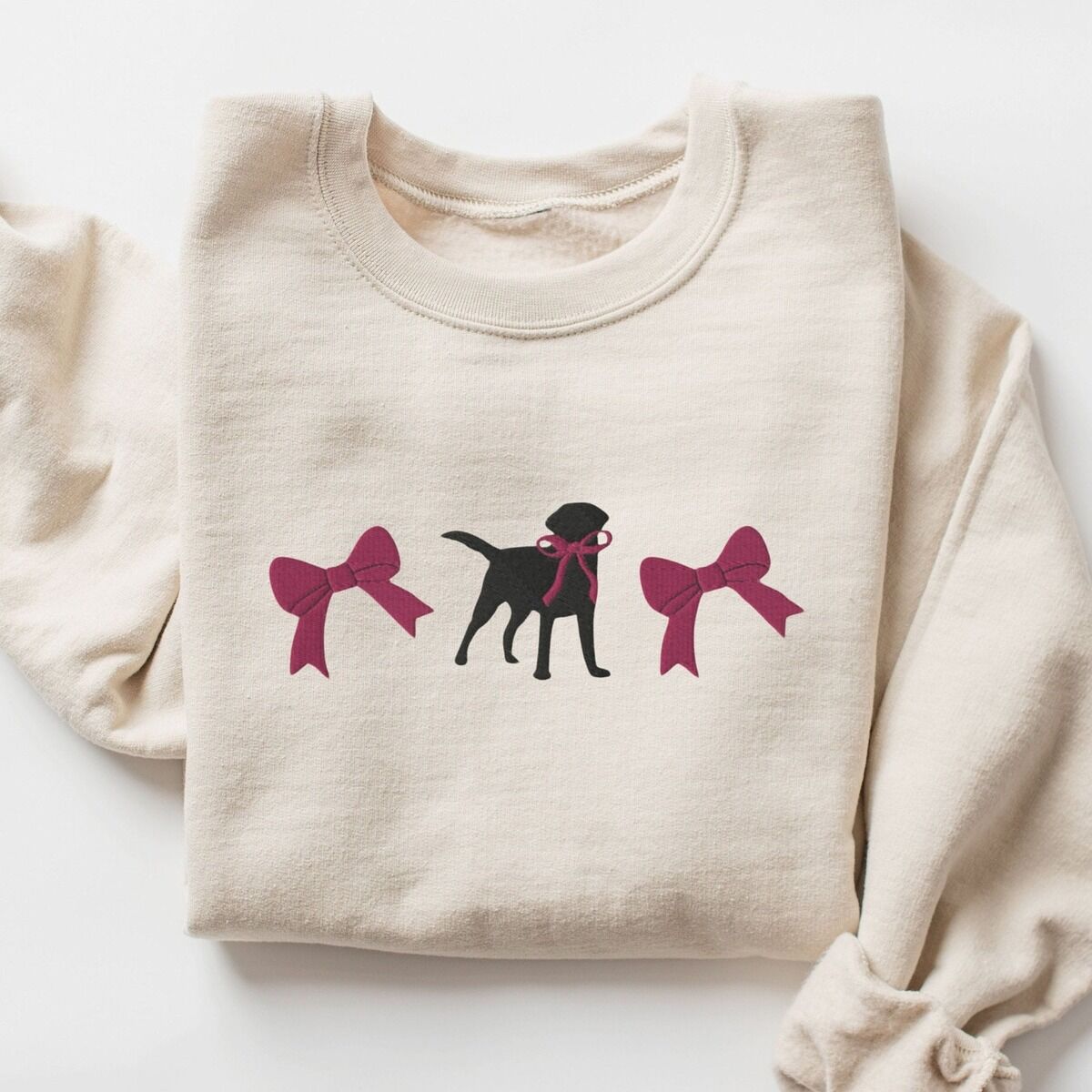 Black Lab Bow Sweatshirt – Sand Sand crewneck sweatshirt laid flat on white, embroidered with a black Labrador silhouette with a ribbon bow at its neck between two large raspberry pink bows across the chest