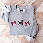 Sport Grey crewneck sweatshirt laid flat on herringbone wood, embroidered with a black Labrador silhouette with a ribbon bow at its neck between two large raspberry pink bows across the chest