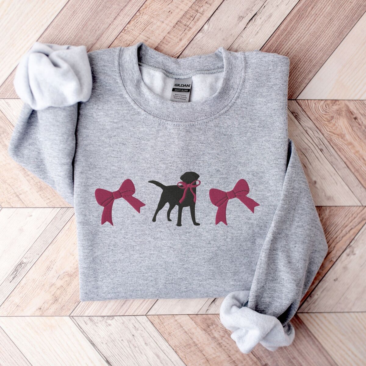 Black Lab Bow Sweatshirt - Sport Grey Sport Grey crewneck sweatshirt laid flat on herringbone wood, embroidered with a black Labrador silhouette with a ribbon bow at its neck between two large raspberry pink bows across the chest