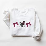 White crewneck sweatshirt laid flat on white, embroidered with a black Labrador silhouette with a ribbon bow at its neck between two large raspberry pink bows across the chest