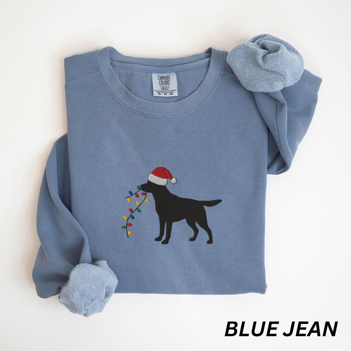 Black Lab Christmas Lights Sweatshirt – Blue Jean Blue Jean crewneck sweatshirt laid flat with rolled cuffs, embroidered with a black Labrador in a Santa hat carrying a string of Christmas lights