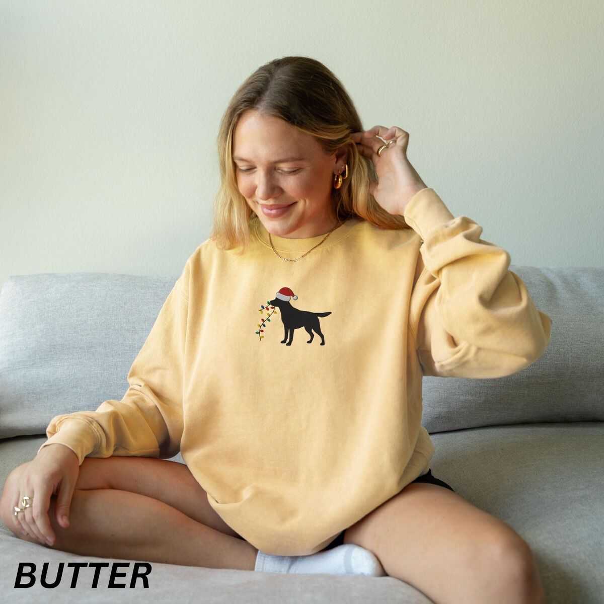 Black Lab Christmas Lights Sweatshirt – Butter Woman on a sofa in a Butter crewneck sweatshirt embroidered with a black Labrador in a Santa hat carrying a string of Christmas lights