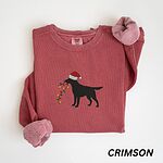 Crimson crewneck sweatshirt laid flat with rolled cuffs, embroidered with a black Labrador in a Santa hat carrying a string of Christmas lights