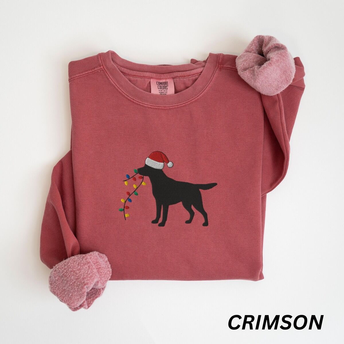 Black Lab Christmas Lights Sweatshirt – Crimson Crimson crewneck sweatshirt laid flat with rolled cuffs, embroidered with a black Labrador in a Santa hat carrying a string of Christmas lights