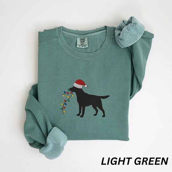 Light Green crewneck sweatshirt laid flat with rolled cuffs, embroidered with a black Labrador in a Santa hat carrying a string of Christmas lights