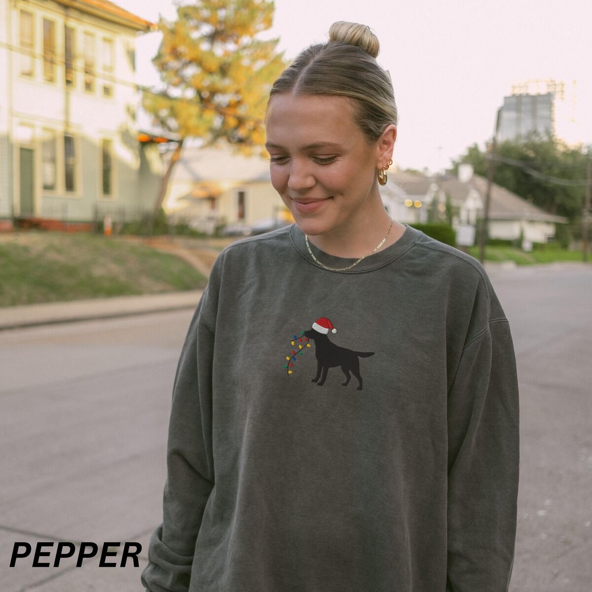 Black Lab Christmas Lights Sweatshirt – Pepper Woman outdoors on a street in a Pepper crewneck sweatshirt embroidered with a black Labrador in a Santa hat carrying a string of Christmas lights