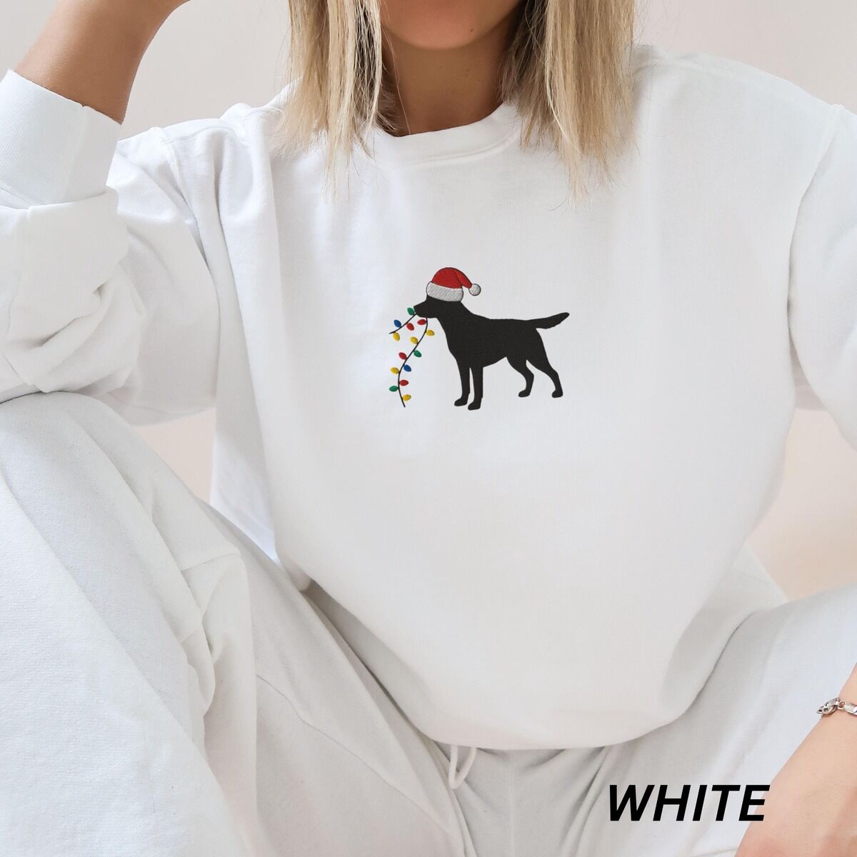 Black Lab Christmas Lights Sweatshirt – White Woman sitting in a White crewneck sweatshirt embroidered with a black Labrador in a Santa hat carrying a string of Christmas lights