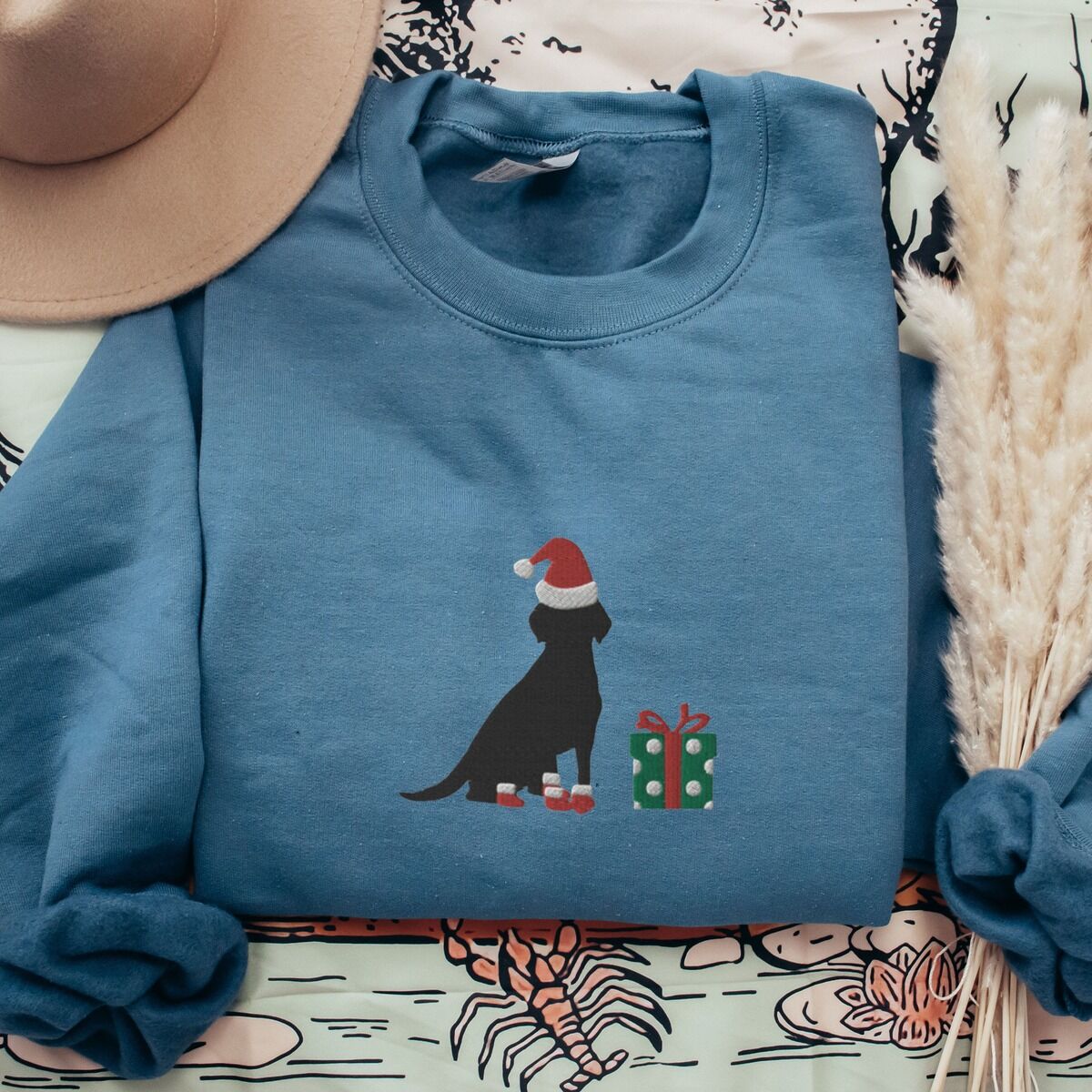 Black Lab Christmas Sweatshirt – Indigo Blue Indigo Blue crewneck sweatshirt laid flat beside a hat and pampas grass, embroidered with a sitting black Labrador in a Santa hat and red booties beside a green gift box